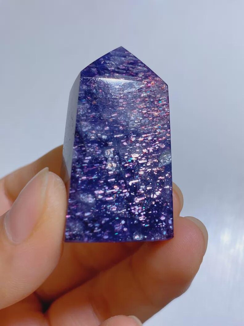 Pu&ograve; includere: Una punta di cristallo lucidata a forma di torre, caratterizzata da un colore di base viola intenso con inclusioni iridescenti simili a glitter che riflettono la luce in tonalit&agrave; rosa, blu e argento sulla sua superficie liscia.