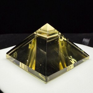 May include: A transparent, yellow-toned pyramid with a square base. The pyramid's edges are sharply defined, and the material appears to be glass or a similar translucent substance. The pyramid is displayed on a white surface against a dark background.