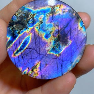 May include: A circular polished labradorite gemstone featuring a vibrant iridescent display of deep purple, electric blue, and golden yellow flashes across its surface, marked by fine natural fissures and dark inclusions throughout the stone.