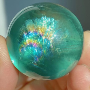 May include: A polished, spherical gemstone with a translucent teal-green body. The interior features a prominent, fan-shaped inclusion that displays a vibrant, iridescent rainbow spectrum of colors, including yellow, blue, and magenta hues.