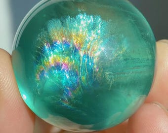 May include: A polished, spherical gemstone with a translucent teal-green body. The interior features a prominent, fan-shaped inclusion that displays a vibrant, iridescent rainbow spectrum of colors, including yellow, blue, and magenta hues.