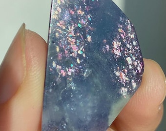 May include: A polished, semi-circular piece of blue stone featuring embedded iridescent flakes that shimmer with pink, gold, and green light. The surface is smooth and translucent, displaying a deep blue base color with scattered reflective inclusions.