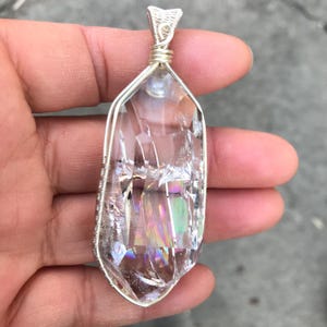 May include: A clear, faceted crystal pendant encased in a silver-colored wire wrap. The crystal features internal fractures that create a rainbow-like iridescent effect when light hits the surface. The wire is coiled at the top to form a bail.