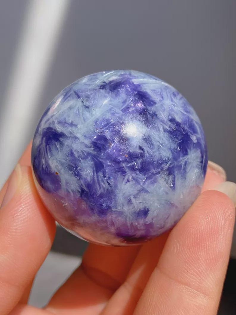 May include: A polished sphere of purple and white crystal. The crystal ball has a marbled appearance with a mix of deep purple and white crystalline structures. The sphere is held in a hand, with a soft, blurred background.