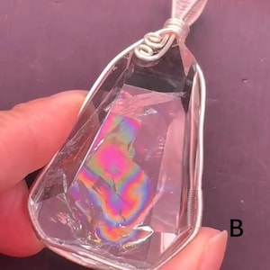 May include: A clear, faceted crystal pendant encased in a silver-colored wire wrap with a decorative spiral bail at the top. The interior of the crystal displays a vibrant, iridescent rainbow-colored inclusion. The letter B is visible on the right.