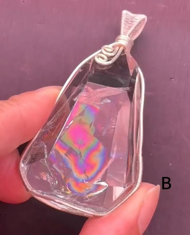 May include: A clear, faceted crystal pendant encased in a silver-colored wire wrap with a decorative spiral bail at the top. The interior of the crystal displays a vibrant, iridescent rainbow-colored inclusion. The letter B is visible on the right.