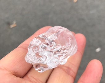Op de afbeelding: Een helder, gebeeldhouwd kristallen beeldje van een mythisch wezen, mogelijk een leeuw of draak. De sculptuur wordt in een hand gehouden en toont zijn ingewikkelde details en doorschijnende kwaliteit. De gravure heeft gedetailleerde gelaatstrekken en een vloeiende manen.
