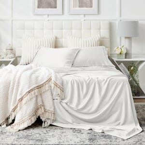 May include: A bed with white sheets, pillows, and a fluffy blanket with fringe. The bed has a white headboard and is next to a white nightstand with a lamp and flowers. The room has a neutral color scheme.