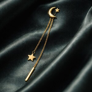 May include: A gold-colored earring with a celestial design. The earring features a crescent moon and star at the top, with a chain that dangles a star and a bar. The earring is set against a dark, silky background.