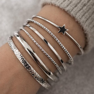 May include: A collection of silver bracelets on a wrist. The bracelets vary in design, including a hammered texture, smooth bands, and a band with small stones. One bracelet features a black star charm.