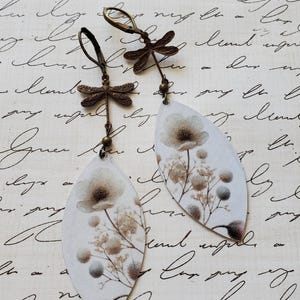 May include: A pair of dangle earrings featuring a botanical design. The earrings have a white background with a floral print in shades of brown and gray. Each earring is leaf-shaped and adorned with a dragonfly charm.