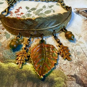 Puede incluir: Una pulsera de metal color latón con una pluma grande y curva como pieza central. De la pluma cuelgan dos pequeños dijes de bellota verde, dos dijes de hoja de roble y un dije central grande con esmalte de color naranja y verde.