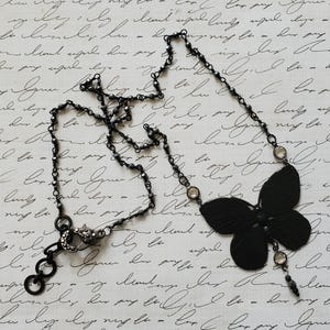 Puede incluir: Un collar negro con un colgante de mariposa. El collar tiene una cadena delicada con peque&ntilde;as cuentas oscuras y detalles redondos transparentes. El colgante de mariposa es de color negro s&oacute;lido.
