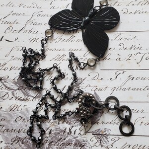 Puede incluir: Collar con colgante de mariposa negra con cadena de cuentas y detalles de cristal transparente. El collar presenta un colgante de mariposa grande y texturizado y un cierre decorativo. El fondo es un papel vintage con escritura.