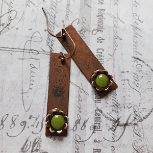 Puede incluir: Pendientes rectangulares de color cobre con un engaste en forma de flor y una piedra verde. Los pendientes tienen un aspecto rústico y envejecido. El fondo presenta escritura y sellos antiguos. Miden unos 4 cm.