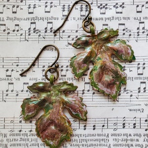 May include: A pair of green and brown leaf-shaped earrings with a gold-toned metal hook. The earrings are on a sheet of music.