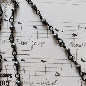Puede incluir: Primer plano de un collar de cuentas negras con cuentas facetadas de color gris oscuro. El collar est&aacute; ensartado en una delicada cadena de alambre negro. El fondo es una hoja de m&uacute;sica vintage con texto manuscrito.