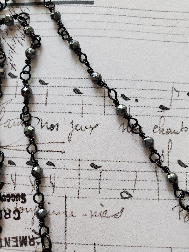 Puede incluir: Primer plano de un collar de cuentas negras con cuentas facetadas de color gris oscuro. El collar est&aacute; ensartado en una delicada cadena de alambre negro. El fondo es una hoja de m&uacute;sica vintage con texto manuscrito.