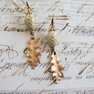 May include: A pair of gold-colored earrings featuring a detailed acorn-shaped bead and a stylized oak leaf charm. The earrings have a gold-tone hook closure. The background is a vintage paper with handwritten script.