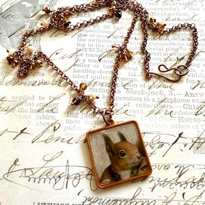 Puede incluir: Un collar de tono cobre con un colgante cuadrado que presenta una imagen detallada de una ardilla roja. La cadena está adornada con pequeñas cuentas de color marrón y crema, y se asegura con un cierre sencillo de gancho.