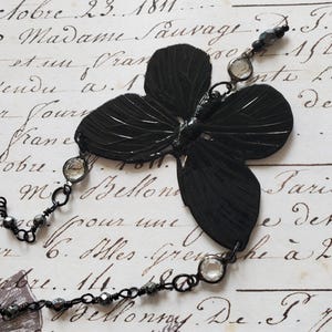 Puede incluir: Un collar con colgante de mariposa negra con un dise&ntilde;o detallado y texturizado. El colgante est&aacute; conectado a una delicada cadena con cuentas transparentes y negras. El collar est&aacute; sobre un fondo de escritura antigua.