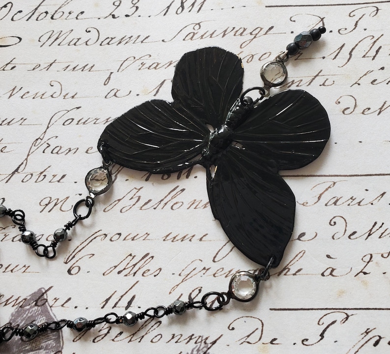 Puede incluir: Un collar con colgante de mariposa negra con un dise&ntilde;o detallado y texturizado. El colgante est&aacute; conectado a una delicada cadena con cuentas transparentes y negras. El collar est&aacute; sobre un fondo de escritura antigua.