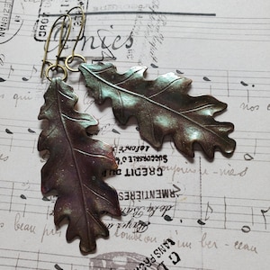 Puede incluir: Pendientes con forma de hoja de roble, con acabado metálico bronce e iridiscente. Los pendientes están suspendidos de ganchos dorados. El fondo es un papel vintage con notas musicales y texto francés.