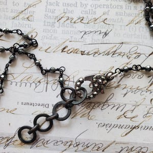 Puede incluir: Primer plano de un collar gris oscuro con un cierre decorativo y m&uacute;ltiples anillos peque&ntilde;os y enlazados. El collar presenta peque&ntilde;as cuentas facetadas ensartadas en alambre. El fondo es un papel vintage con texto manuscrito.
