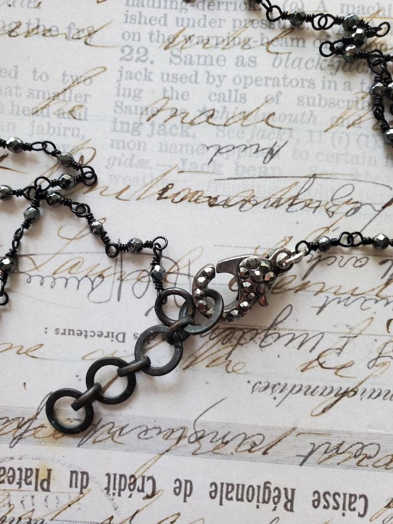 Puede incluir: Primer plano de un collar gris oscuro con un cierre decorativo y m&uacute;ltiples anillos peque&ntilde;os y enlazados. El collar presenta peque&ntilde;as cuentas facetadas ensartadas en alambre. El fondo es un papel vintage con texto manuscrito.