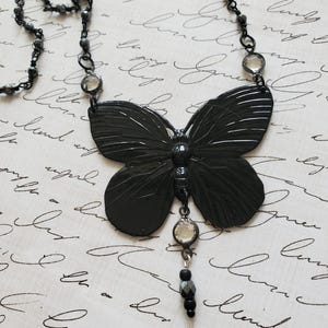 Puede incluir: Un collar con colgante de mariposa negra. El collar presenta un colgante de mariposa grande y detallado con una cadena de cuentas y detalles de cristal transparente. La mariposa tiene alas texturizadas y un peque&ntilde;o dije colgante.