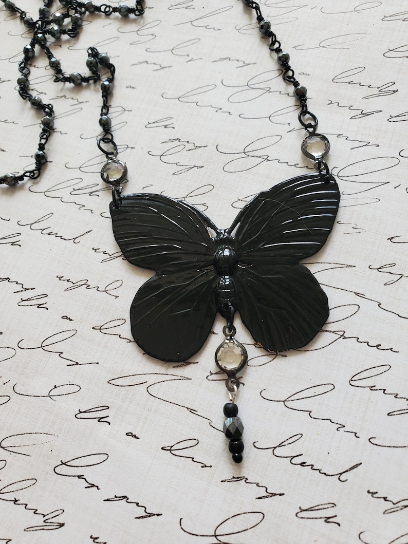 Puede incluir: Un collar con colgante de mariposa negra. El collar presenta un colgante de mariposa grande y detallado con una cadena de cuentas y detalles de cristal transparente. La mariposa tiene alas texturizadas y un peque&ntilde;o dije colgante.