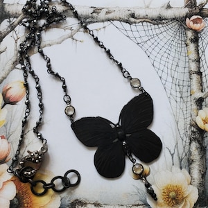 Puede incluir: Un collar con colgante de mariposa negra con una cadena de cuentas y detalles de cristal transparente. La mariposa tiene patrones de alas detallados. El collar tiene un cierre de mosquet&oacute;n.