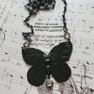Puede incluir: Un collar con colgante de mariposa gris oscuro. El collar presenta un colgante de mariposa grande y detallado con una cadena de cuentas y detalles. El fondo es un papel vintage con notas musicales y texto en franc&eacute;s.