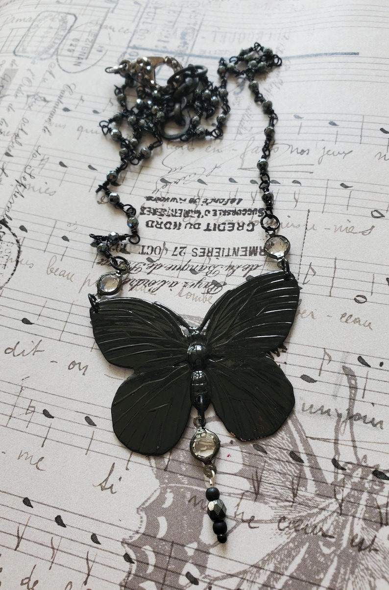 Puede incluir: Un collar con colgante de mariposa gris oscuro. El collar presenta un colgante de mariposa grande y detallado con una cadena de cuentas y detalles. El fondo es un papel vintage con notas musicales y texto en franc&eacute;s.