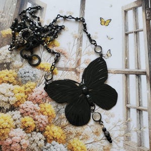 Puede incluir: Un collar con colgante de mariposa gris oscuro. El colgante de mariposa es el punto focal, con detalles intrincados. El collar presenta una delicada cadena con peque&ntilde;as cuentas grises oscuras y acentos transparentes. El fondo incluye un dise&ntilde;o floral y una ventana.