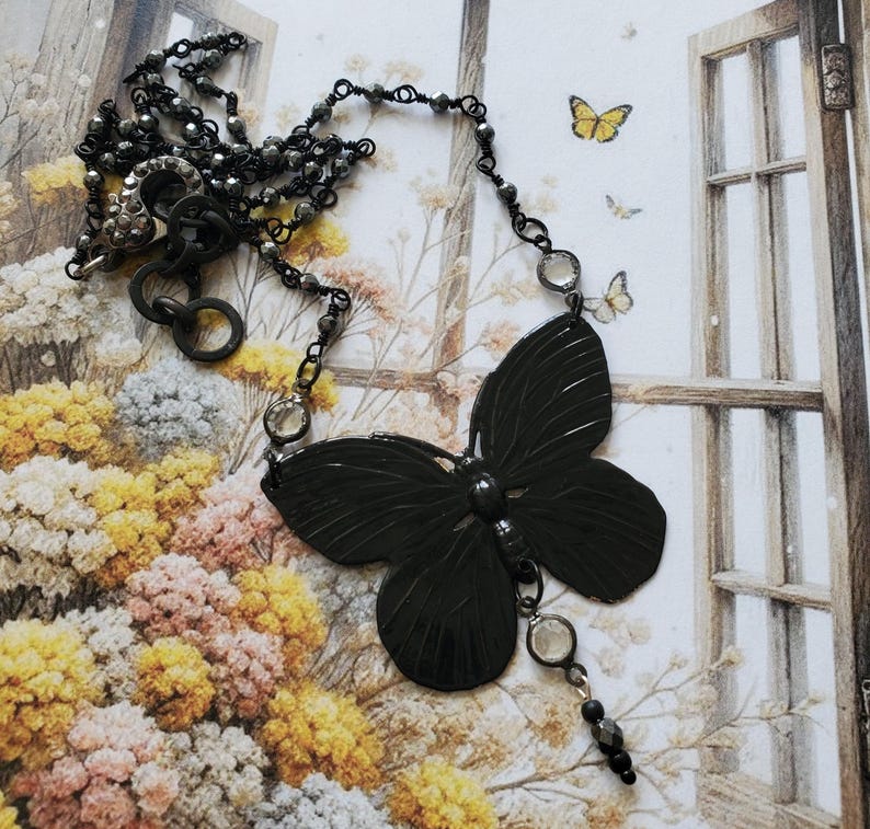 Puede incluir: Un collar con colgante de mariposa gris oscuro. El colgante de mariposa es el punto focal, con detalles intrincados. El collar presenta una delicada cadena con peque&ntilde;as cuentas grises oscuras y acentos transparentes. El fondo incluye un dise&ntilde;o floral y una ventana.