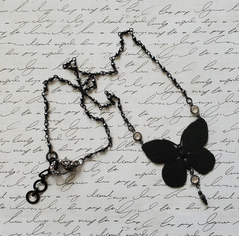 Puede incluir: Un collar negro con un colgante de mariposa. El collar tiene una cadena delicada con peque&ntilde;as cuentas oscuras y detalles redondos transparentes. El colgante de mariposa es de color negro s&oacute;lido.