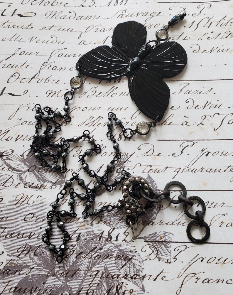 Puede incluir: Collar con colgante de mariposa negra con cadena de cuentas y detalles de cristal transparente. El collar presenta un colgante de mariposa grande y texturizado y un cierre decorativo. El fondo es un papel vintage con escritura.