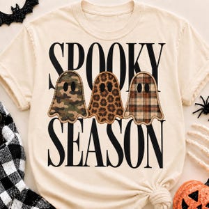 Op de afbeelding: Een crèmekleurig t-shirt met de zwarte tekst "SPOOKY SEASON" en drie geestvormige afbeeldingen. De geesten hebben verschillende patronen: één met groene camouflage, één met een bruine luipaardprint en één met een bruin en zwart ruitpatroon.