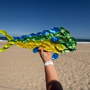 May include: A decorative fish sculpture crafted from numerous individual oyster shells painted in vibrant shades of yellow, green, and blue. The shells are arranged to form the elongated, streamlined body shape of a mahi-mahi or dorado fish.