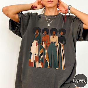 May include: Dark gray t-shirt featuring a graphic of four women with afros, each in a different outfit. The outfits include a variety of colors and patterns, including skirts, pants, and tops. The word "PEPPER" is in a circle on the bottom right.