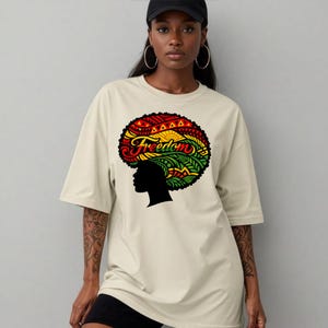 May include: Beige t-shirt featuring a colorful graphic of an afro with the word "Freedom" in red and green. The graphic includes red, yellow, and green accents. The shirt has short sleeves and a relaxed fit.