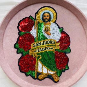 Könnte beinhalten: Gestickter Aufnäher mit San Judas Tadeo, umgeben von roten Paillettenrosen. Der Heilige ist in grünen und weißen Gewändern dargestellt, mit goldenem Heiligenschein und Stab. Das Banner lautet "SAN JUDAS TADEO".