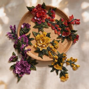 Può includere: Tre toppe ricamate a motivi floreali con intricate perline e paillettes in vivaci tonalità di rosso, giallo e viola. Ogni elemento mostra foglie e steli verdi dettagliati, caratterizzati da un design a petali sovrapposti con riflessi.