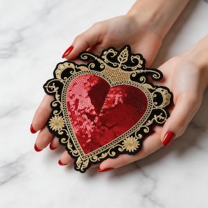 May include: A heart-shaped patch with red sequins, framed by black and gold embroidery. The patch features a crown design at the top and floral accents. The patch is held in two hands with red nail polish.
