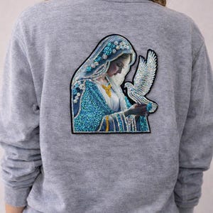 Op de afbeelding: Een grijze sweatshirt met een gedetailleerde patch van een vrouw in een blauw-witte versierde jurk, die een witte duif vasthoudt. De patch heeft een zwarte rand. De vrouw draagt een blauwe hoofdbedekking.