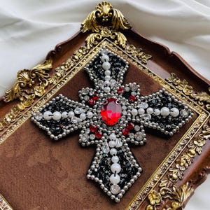 May include: Ornate cross with a large red gemstone center, surrounded by black and silver beads, white pearls, and smaller red accents. The cross is set on a brown velvet background within a decorative, gold-trimmed frame.