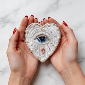 May include: Heart-shaped white embroidered brooch with a blue eye design. The brooch is decorated with pearls and sequins, and has a small pink crystal. The brooch is held in two hands.