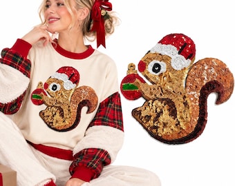 Puede incluir: Una sudadera color crema con una gran aplicación de lentejuelas de una ardilla con gorro de Papá Noel que sostiene un adorno navideño. Las mangas tienen paneles de tela de cuadros rojos y verdes y secciones de forro polar blanco.