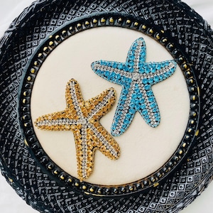 Könnte beinhalten: Zwei mit Juwelen besetzte Seesterne, einer goldfarben und einer türkis, werden auf einer cremefarbenen Oberfläche in einem dekorativen schwarzen Rahmen präsentiert. Die Seesterne sind mit glitzernden Kristallen bedeckt.